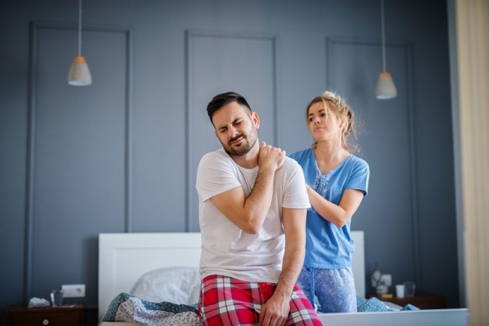 Tired unhappy man holding his neck after bad sleep while his wife looking sadly to him and helping with massage.